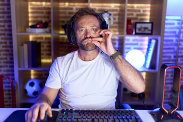 Middle age man with beard playing video games wearing headphones mouth and lips shut as zip with fingers. secret and silent, taboo talking