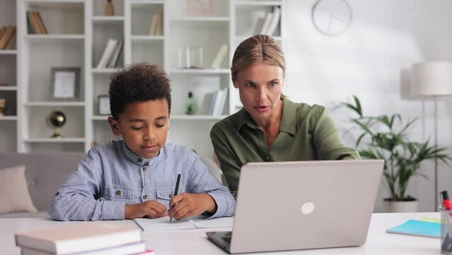 Caring female tutor helping little African American elementary school student with homework at home while sitting at the table. Happy boy doing homework on laptop, studying at home with babysitter.