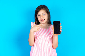 Smiling caucaisna kid girl wearing pink dress over blue background Mock up copy space. Pointing index finger on mobile phone with blank empty screen