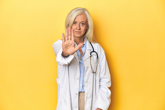 Middle-aged Caucasian female doctor on yellow studio backdrop Middle-aged Caucasian female doctor on yellow studio backdropstanding with outstretched hand showing stop sign, preventing you.