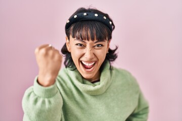 Young beautiful woman standing over pink background angry and mad raising fist frustrated and furious while shouting with anger. rage and aggressive concept.