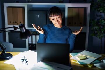 Young beautiful woman working at the office at night clueless and confused expression with arms and...
