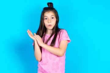 Surprised emotional Caucasian kid girl wearing pink t-shirt over blue background rubs palms and stares at camera with disbelief