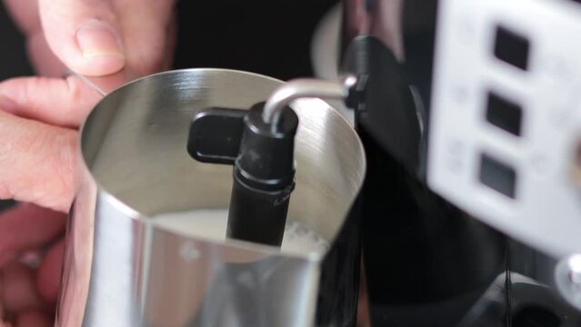 A barista froths milk in a metal mug using a cappuccino maker in a coffee maker. Close-up of a mug with milk. Morning coffee.