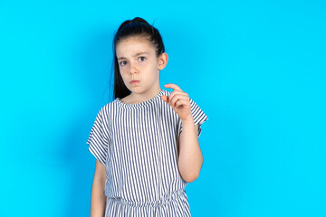 Displeased Caucasian kid girl wearing striped dress over blue background shapes little hand sign demonstrates something not very big. Body language concept.