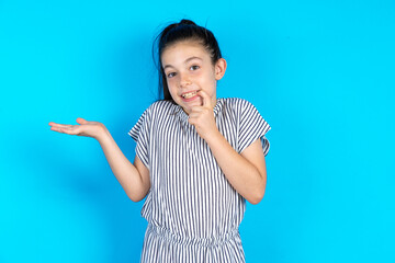 Positive Caucasian kid girl wearing striped dress over blue background advert promo touch finger teeth