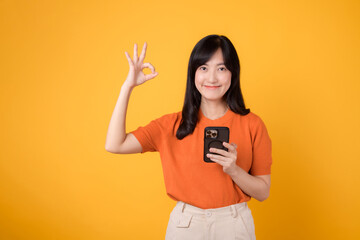 Cheerful Asian woman in her 30s, wearing orange shirt, using smartphone with okay hand sign on...