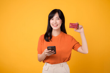 Excited Asian woman 30s in orange shirt, using smartphone and showing credit card on yellow background. Fast online shopping.