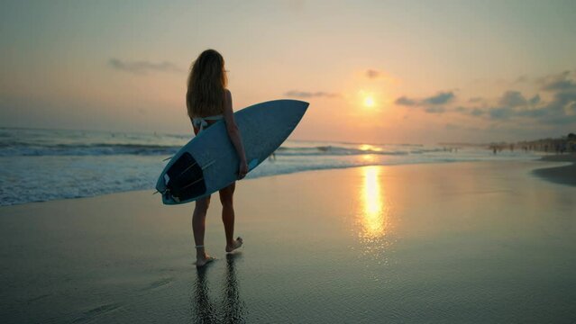 Tourist woman surfer in swimsuit with surfboard stands on ocean sandy beach at sunset admires waves. Summer sport activity on tropics resort. Sporty slim girl. Extreme water sport surfing on vacation.