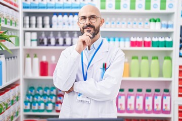 Hispanic man with tattoos working at pharmacy drugstore smiling looking confident at the camera with crossed arms and hand on chin. thinking positive.