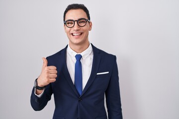 Young hispanic man wearing suit and tie doing happy thumbs up gesture with hand. approving expression looking at the camera showing success.