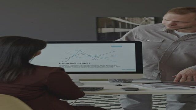 Vertical medium shot of businesswoman sitting at work desk with graph of company financial progress on computer monitor, discussing indicators with male office worker