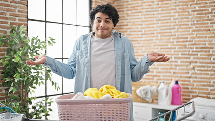 Young latin man standing by basket with clothes clueless at laundry room