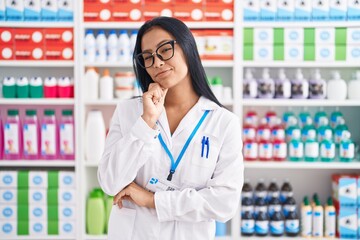 Hispanic woman working at pharmacy drugstore serious face thinking about question with hand on chin, thoughtful about confusing idea