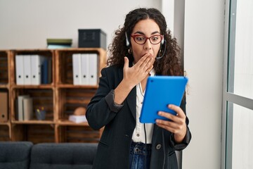Young hispanic woman using touchpad wearing headphones covering mouth with hand, shocked and afraid...
