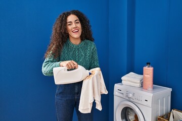 Young hispanic woman holding dirty laundry and detergent bottle smiling and laughing hard out loud because funny crazy joke.