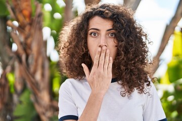 Hispanic woman with curly hair standing outdoors covering mouth with hand, shocked and afraid for...