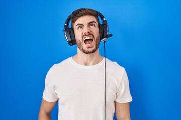 Hispanic man with beard listening to music wearing headphones angry and mad screaming frustrated and furious, shouting with anger. rage and aggressive concept.
