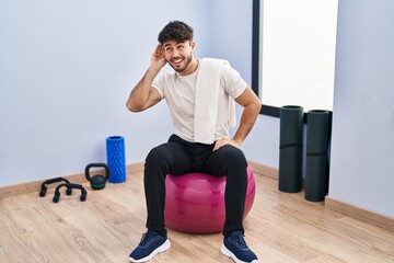 Hispanic man with beard sitting on pilate balls at yoga room smiling with hand over ear listening an hearing to rumor or gossip. deafness concept.