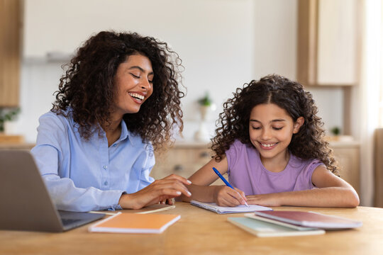 Loving latin mother working on laptop and helping her daughter with study, family doing homework together, sitting at table in living room interior