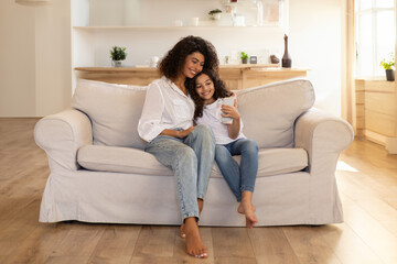 Fototapeta premium Adorable latin mother and daughter using cellphone, sitting on sofa in living room, spending time together at home, having video chat, copy space