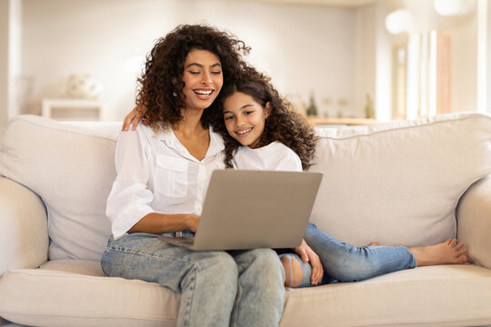 Family digital leisure. Cheerful latin mother and daughter using laptop engaging in fun online activities, surfing internet or video calling sitting on sofa at home, free space