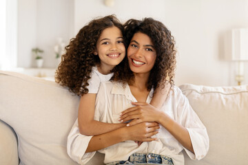 Happy latin girl hugging her beautiful mother and smiling together at camera, sitting on sofa in living room, adorable family embracing at home - obrazy, fototapety, plakaty