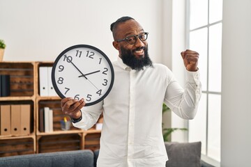 African american man holding big clock at the office screaming proud, celebrating victory and success very excited with raised arm