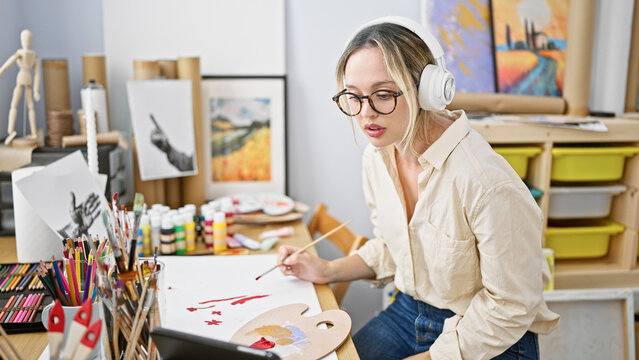 Young blonde woman artist drawing on paper using touchpad and headphones at art studio