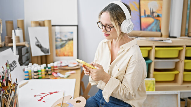 Young blonde woman artist sitting on table using smartphone and headphones at art studio
