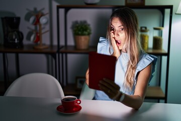 Young hispanic woman using touchpad sitting on the table at night afraid and shocked, surprise and...