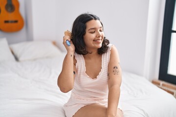 Young woman combing hair sitting on bed at bedroom