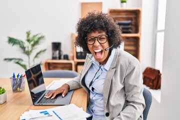 Black woman with curly hair wearing call center agent headset at the office angry and mad screaming...