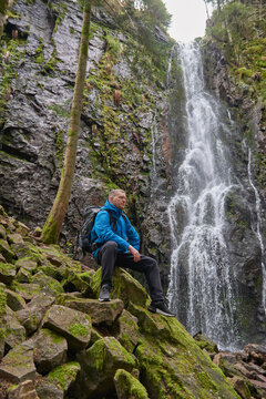 Tourist attraction of Germany - falls of Burgbach Waterfall near Schapbach, Black Forest, Baden-Wurttemberg, Germany. Man hiker in blue jacket standing on stone and looks at flow of falling water
