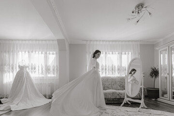 Happy dark-haired brunette bride in a satin robe and professional make-up standing in her room near the mirror and getting ready for her wedding day