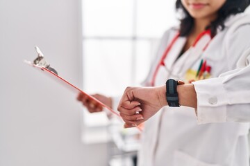 Young latin woman doctor reading document looking watch at clinic