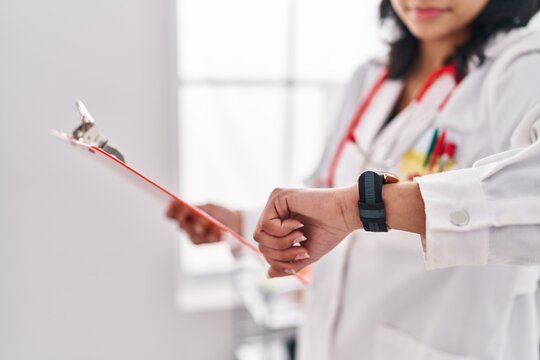 Young Latin Woman Doctor Reading Document Looking Watch At Clinic