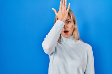 Young caucasian woman standing over blue background surprised with hand on head for mistake,...