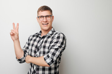 Portrait of young happy caucasian man in shirt showing peace and love gesture with fingers isolated on white studio background