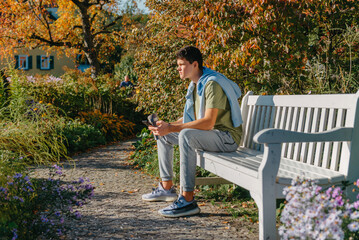 Fototapeta premium A Teenager Sits On A Bench In The Autumn Park Drinks Coffee From A Thermo Mug And Looks Into A Phone. Portrait Of Handsome Cheerful Guy Sitting On Bench Fresh Air Using Device Browsing Media Smm
