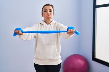 Middle age hispanic woman training arm resistance with elastic arm bands puffing cheeks with funny face. mouth inflated with air, catching air.