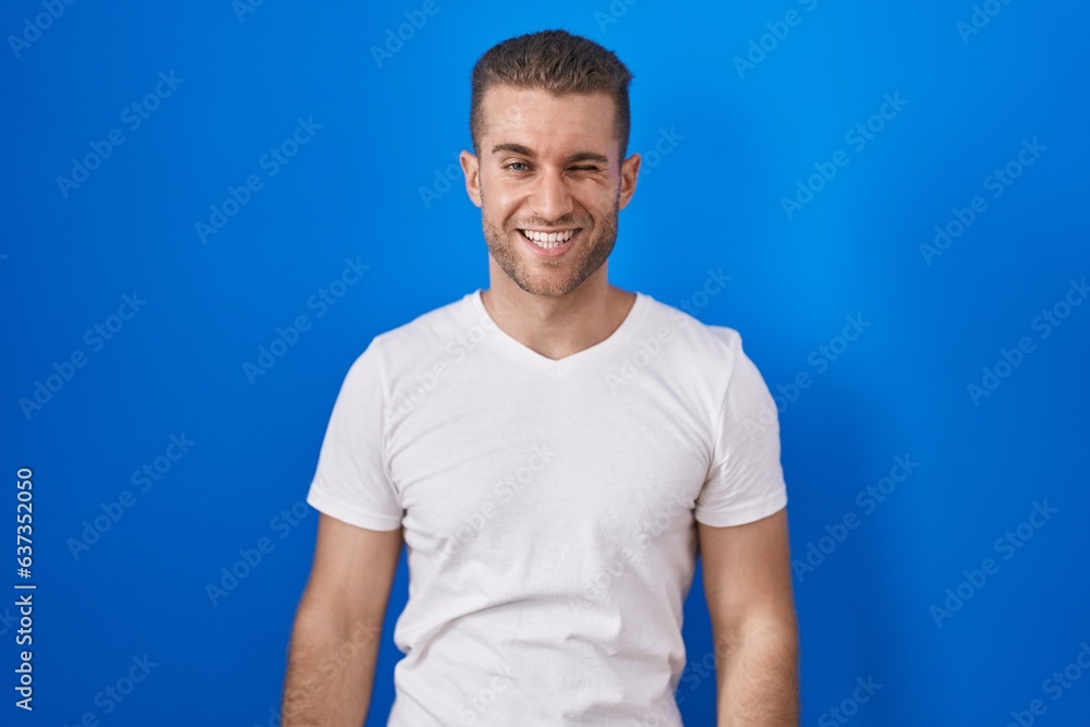 Poster young caucasian man standing over blue background winking looking at the camera with sexy expression