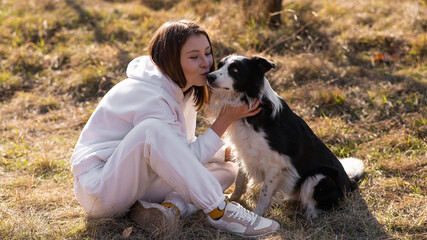Caucasian woman hugging and kissing her Border Collie dog while sitting on grass in autumn park.