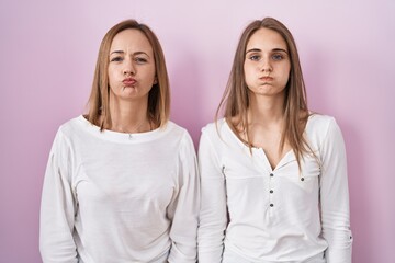 Middle age mother and young daughter standing over pink background puffing cheeks with funny face. mouth inflated with air, crazy expression.