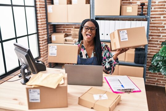African woman with braids working at small business ecommerce holding packages smiling and laughing hard out loud because funny crazy joke.