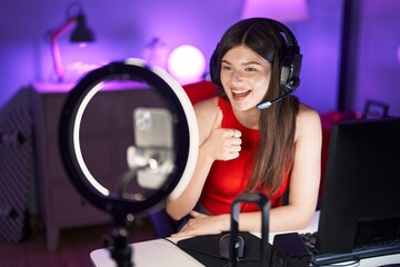 Young caucasian woman playing video games recording with smartphone smiling happy and positive, thumb up doing excellent and approval sign
