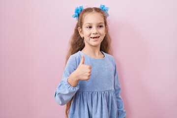 Young little girl standing over pink background smiling happy and positive, thumb up doing excellent and approval sign