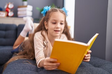 Adorable caucasian girl reading book lying on sofa at home