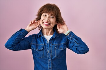 Middle age woman standing over pink background smiling pulling ears with fingers, funny gesture....