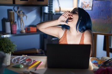 Fototapeta na wymiar Young modern girl with blue hair sitting at art studio with laptop at night shouting and screaming loud to side with hand on mouth. communication concept.